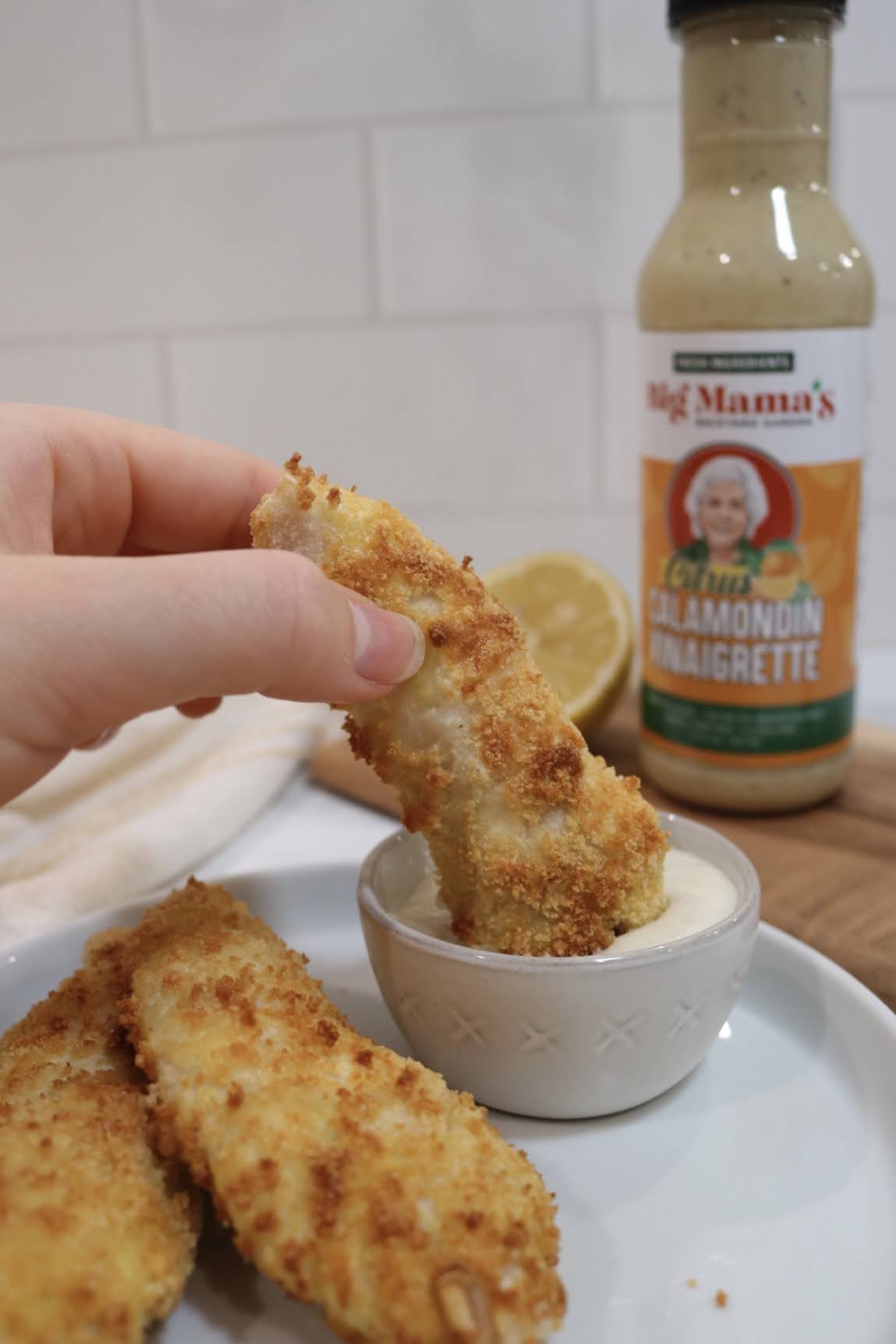 a hand dipping a gluten-free air fryer chicken tender into a dipping sauce in a cup. a bottle of citrus calamondin vinaigrette is in the background.